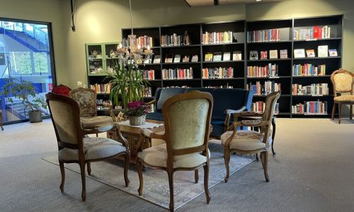 Et koselig bibliotek eller leserom med bokhyller langs bakveggen. I forgrunnen er antikke stoler og en sofa plassert rundt et salongbord. En lysekrone henger over, og det er et stort vindu på venstre side. Rommet har en blanding av moderne og klassiske elementer, som skaper en innbydende atmosfære for lesing eller avslapning.