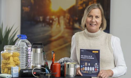 En dame som holder en brosjyre som vises frem. På bordet står det ting man skal ha hjemme i beredskap, som vann, tørrmat, gass.